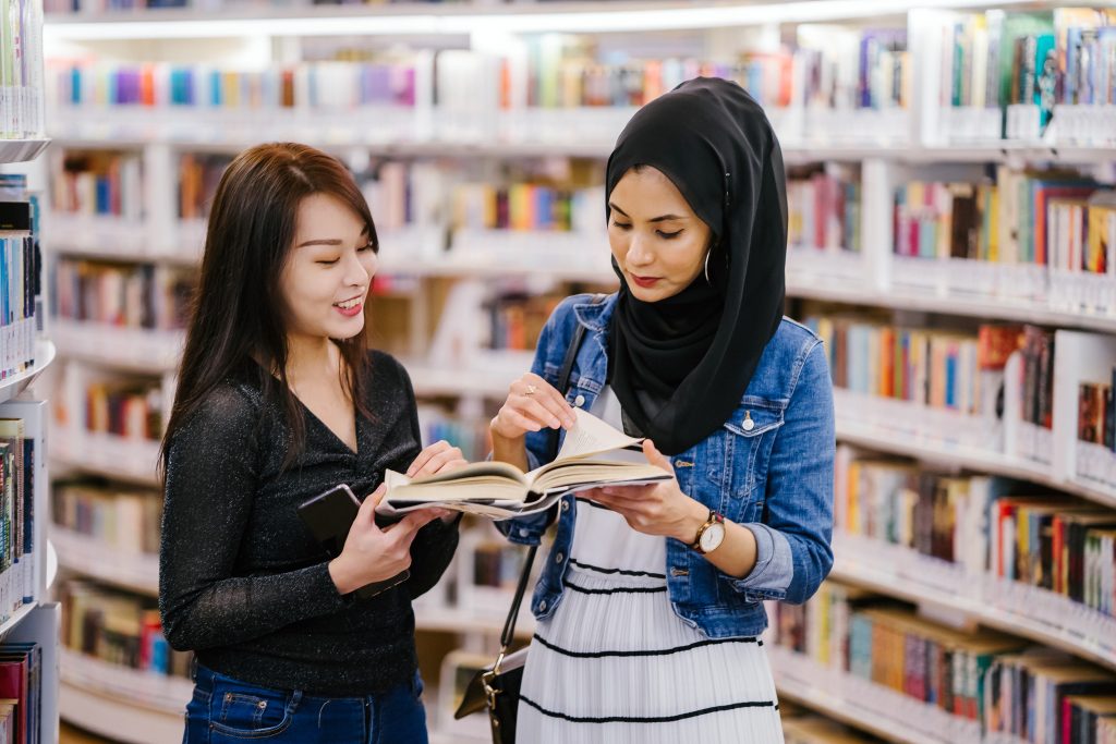 A Chinese Asian woman and a Muslim woman stand and talk together over a book in a library. They are both young, attractive and smiling as they have a productive discussion together. 